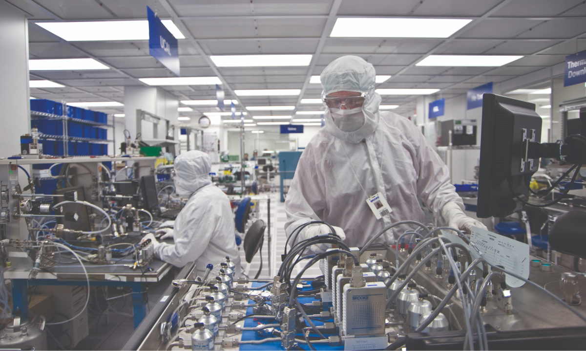 Two Brooks Instrument employees in white suits collaborate on instruments in a clean room setting.
