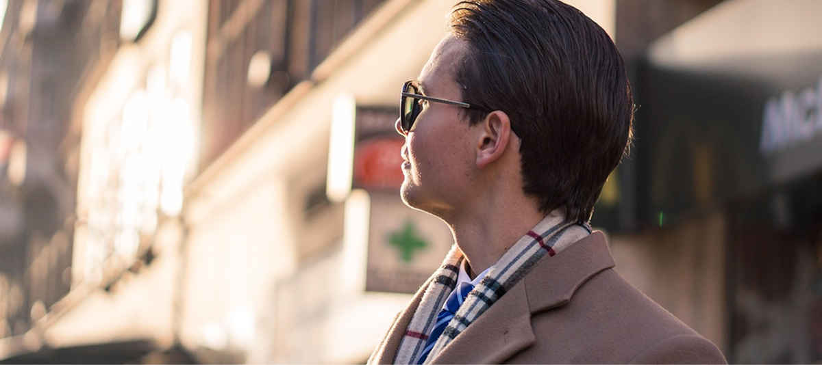 A man dressed in a coat and scarf walks down a street, with shops visible in the background.
