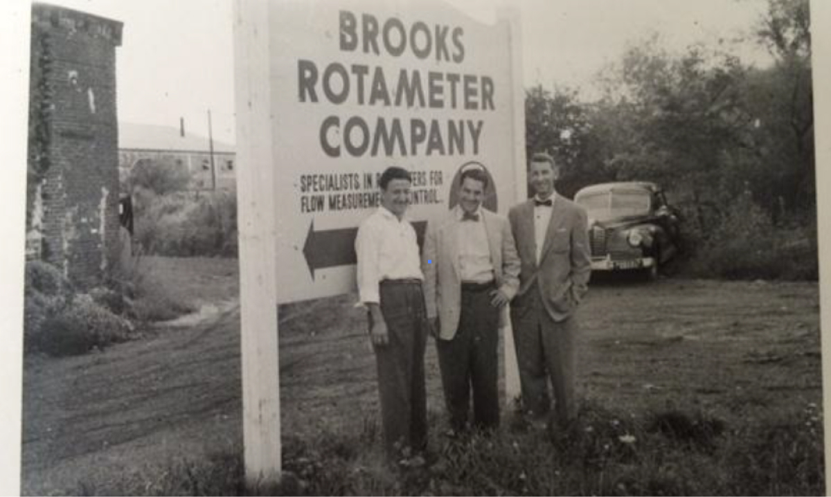 Three men pose in front of a sign for Brooks Rotameter Company, highlighting its historical significance in the industry.