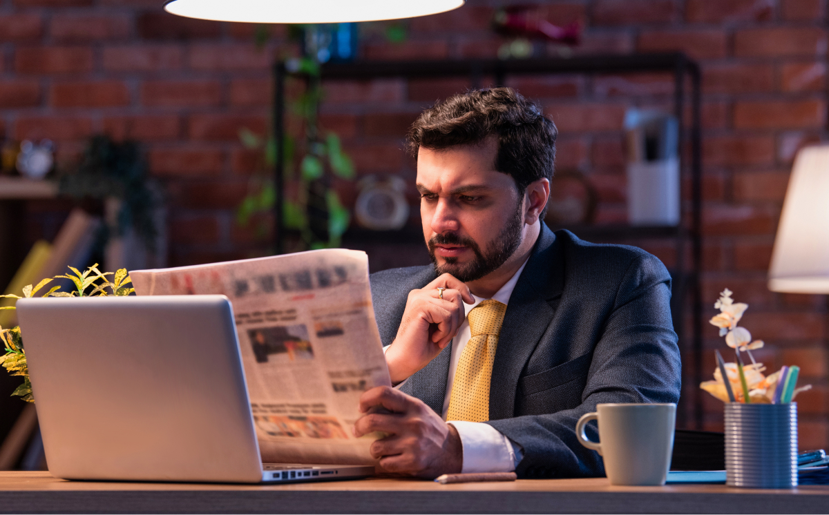A man in a suit reads a newspaper while seated at a desk, focused on the content in front of him.
