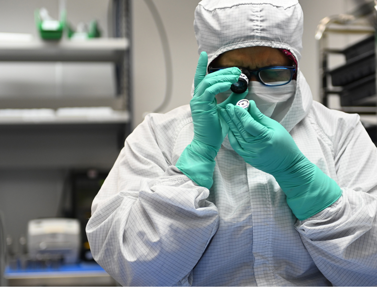 A Brooks Instrument employee in a lab coat and gloves holds a small object, focused on her task in a laboratory setting.