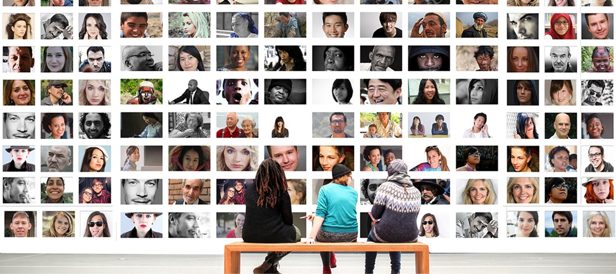 Two people sit on a bench, gazing at a wall filled with various photographs.