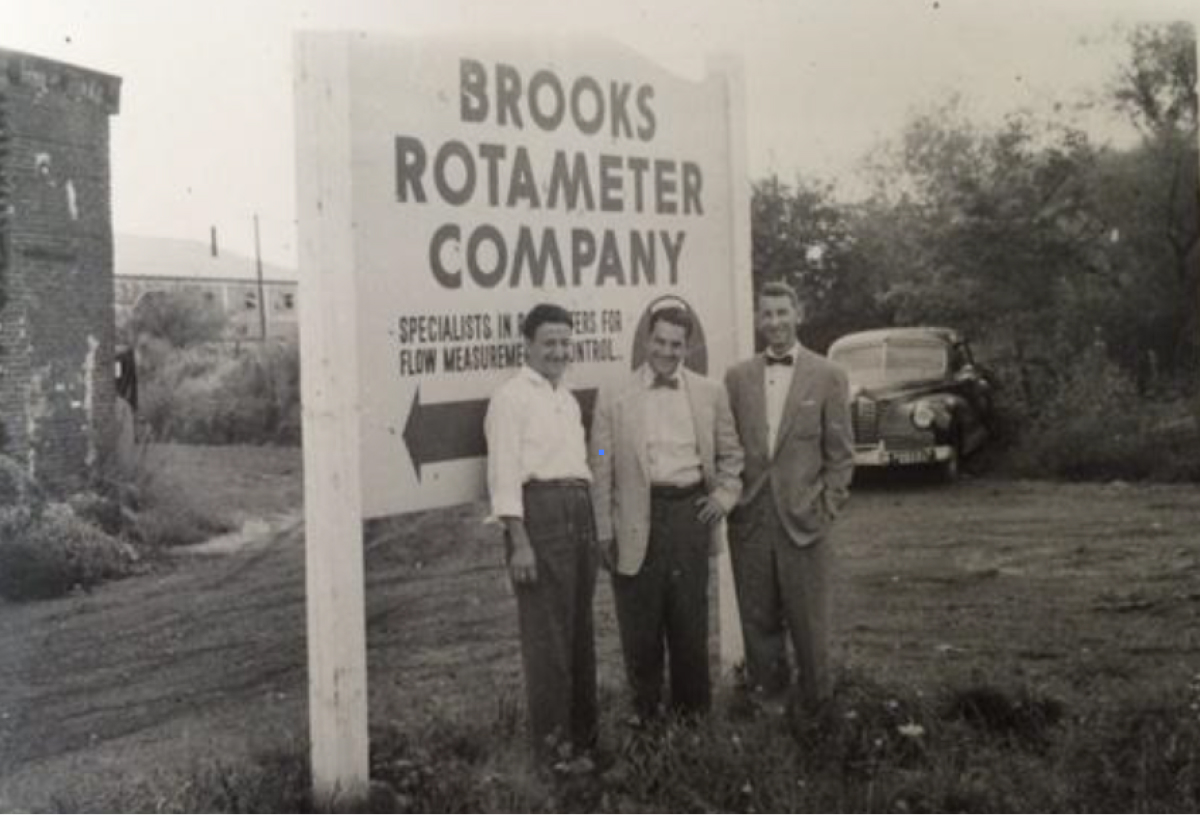 Three men stand in front of a sign for Brooks Rotameter Company, showcasing the company's history and presence.