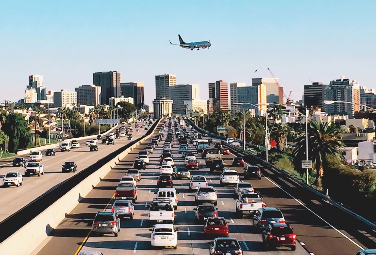A plane soars above a busy freeway filled with cars, with buildings visible in the background.
